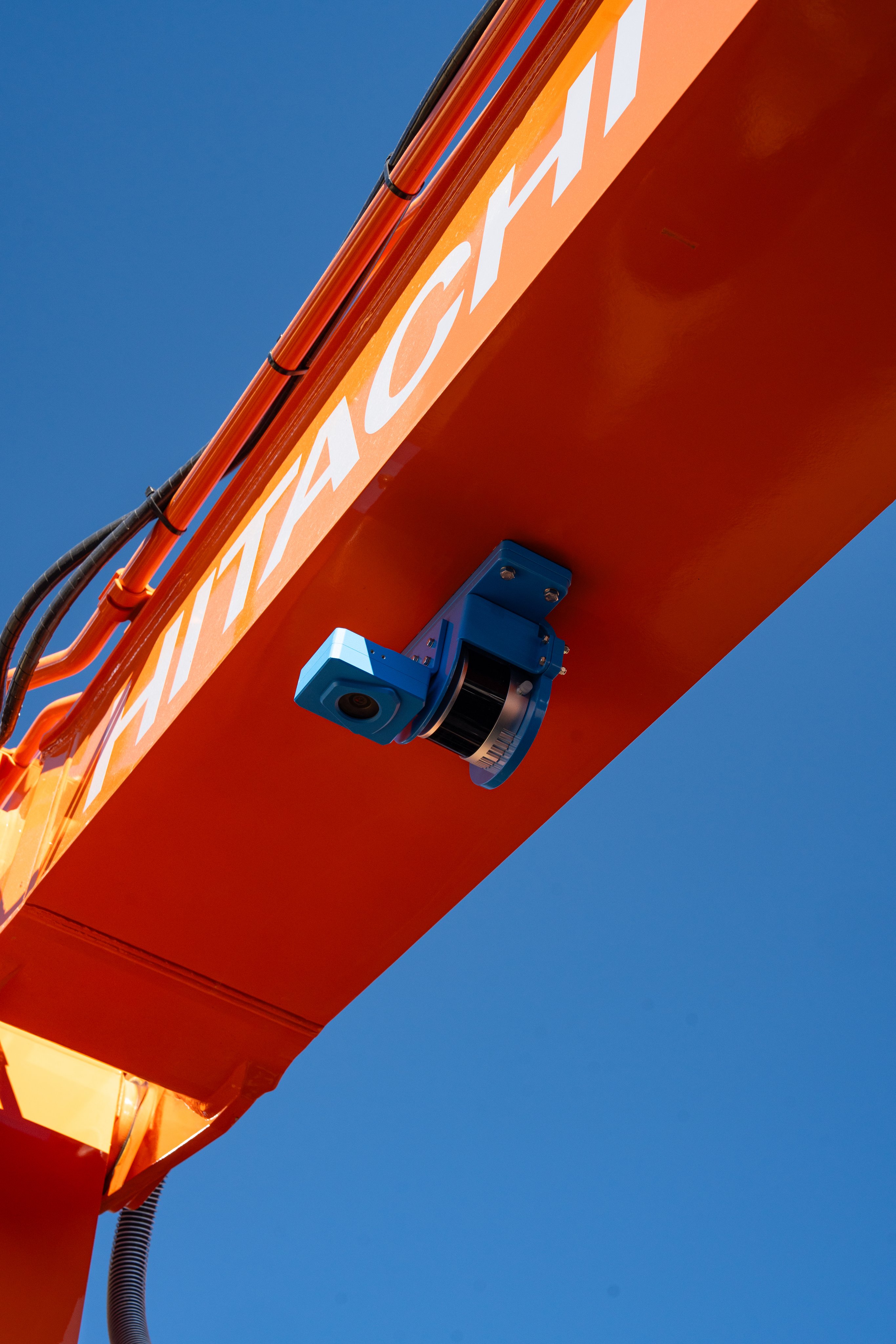 Blue SDX-4DVision sensor housing on the underside of an orange excavator boom against a blue sky